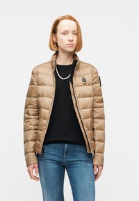 Νεαρή γυναίκα με κοντά κόκκινα μαλλιά, που φοράει μια μπεζ μπουφάν με καπιτονέ σχέδιο, μαύρο τοπ, κολιέ με μαργαριτάρια και μπλε τζιν, στέκεται μπροστά σε απλό φόντο.