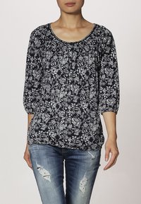 Blusa floral azul marino con mangas de 3/4, de corte holgado, que presenta patrones florales en blanco, escote fruncido y una textura suave. Combinada con jeans desgastados.