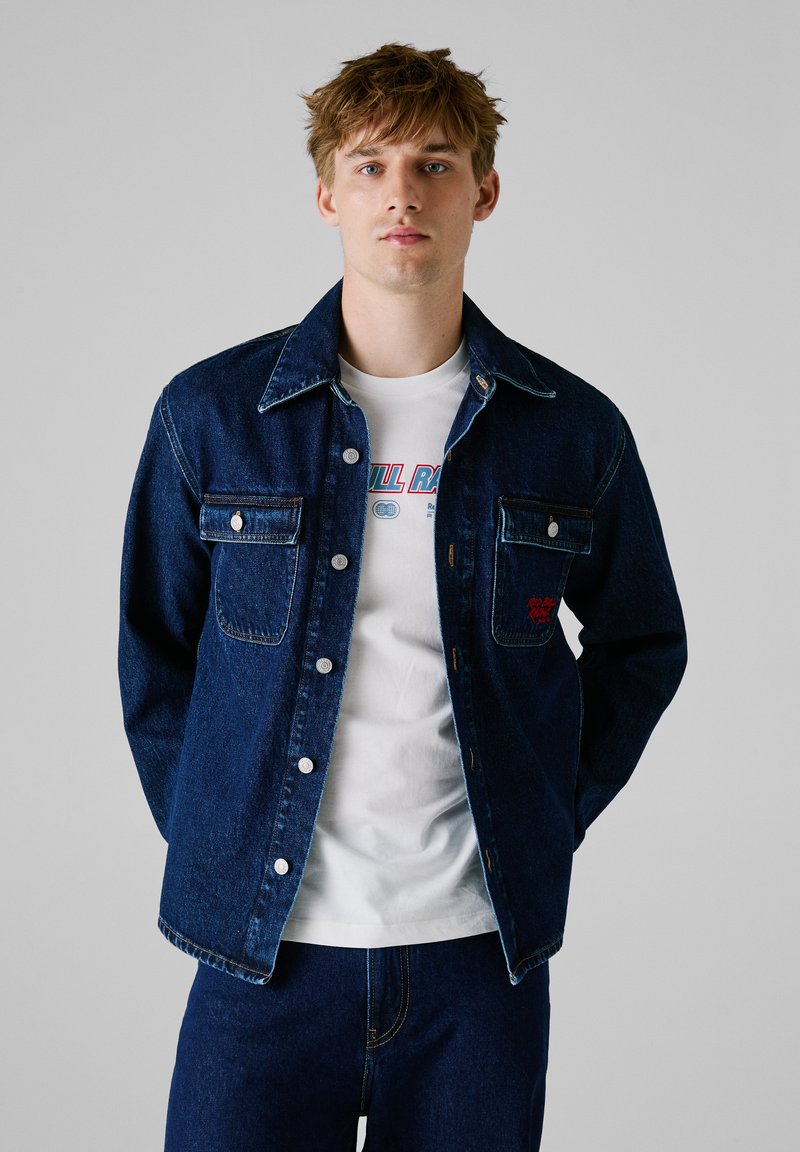 Chaqueta de mezclilla en azul oscuro con botones plateados, dos bolsillos en el pecho, costuras en contraste, llevada sobre una camiseta gráfica blanca. Corte casual.