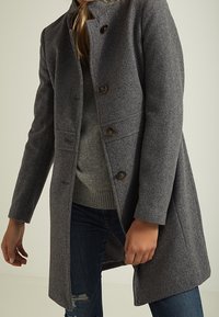 Grå ullkappa med hög krage, sju knappar och fickor. Designen har en skräddarsydd passform och ett något texturerat tyg.