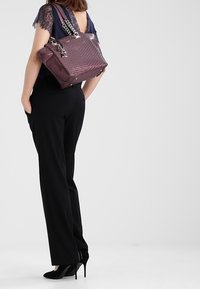 Mujer con blusa de mangas de encaje y pantalones negros, sosteniendo un bolso púrpura elegante, de pie frente a un fondo liso.