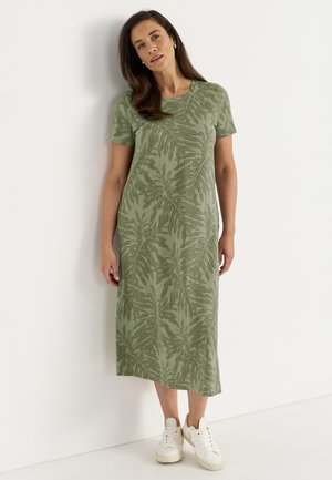 Kvinde iført en grøn midi-kjole med et bladmønster og hvide sneakers, der står op ad en hvid væg.