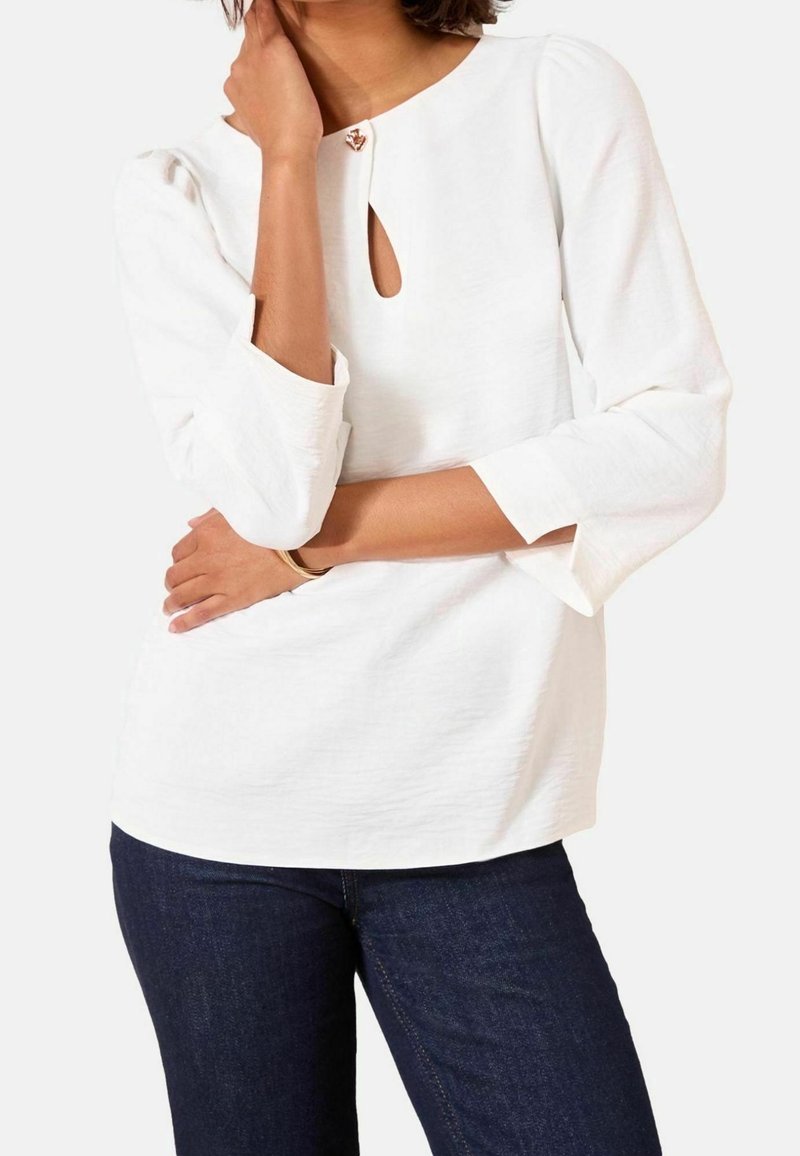 Femme portant un haut blanc à manches longues avec un décolleté en forme de trou de serrure et un jean bleu foncé, les bras croisés et une main touchant son cou.