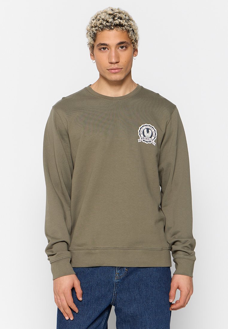 Młoda osoba z krótkimi, kręconymi blond włosami, ubrana w oliwkową koszulę z długim rękawem z logo na piersi oraz niebieskie dżinsy, stojąca na tle białym.