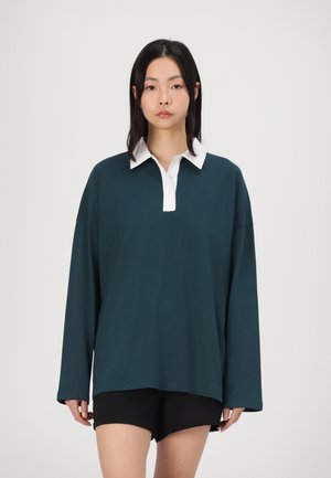 Mørk teal langermet polo skjorte med hvit krage, med en løs passform og mykt stoff. Brukt med svarte shorts.