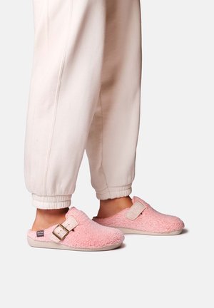 Personne portant des chaussons légers rose pâle à enfiler avec des sangles beige à boucle et un pantalon crème avec revers, sur un fond uni.