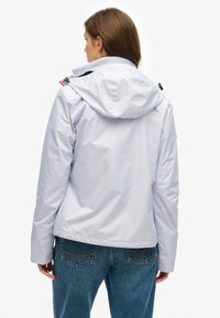 Jasnoszara wodoodporna kurtka z kapturem, z logo na plecach, regulowanym kołnierzem, zestawiona z niebieskimi dżinsami. Gładka faktura.