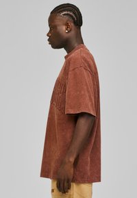 Brun oversized T-skjorte med teksturert finish og preget tekst over fronten. Korte ermer og rund hals-design.