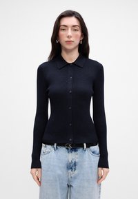 Námornícky pruhovaný pletený kardigán s klasickým golierom, dlhými rukávmi a gombíkovým zapínaním. Nosený cez svetlomodré džínsy s tmavým opaskom.