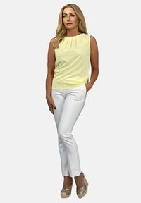 Żółta bezrękawnikowa bluzka z plisowanym dekoltem, zestawiona z białymi dopasowanymi spodniami. Modelka nosi beżowe buty na platformie o strukturalnym wykończeniu.