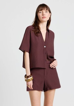 Woman wearing a loose, short-sleeve maroon shirt and matching shorts, accessorized with stacked bangles and rings on left hand.