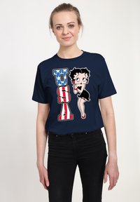 Námořnická modrá trička s ilustrací postavy v černých šatech, s nápisem "USA" ve velkých červených a modrých písmenech, doplněné hvězdami.