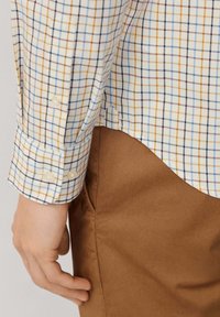 Camisa a cuadros con líneas azules, amarillas y marrones sobre un fondo blanco, con un puño abotonado y una textura suave. Pantalones marrones parcialmente visibles.