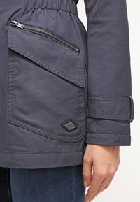 Dark gray jacket featuring a cinched waist, zippered chest pocket, large front flap pocket, and branded tag detail. Textured fabric.