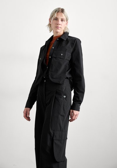 Giacca cropped nera con due tasche sul petto e bottoni argentati, abbinata a pantaloni neri a vita alta con tasche cargo e dettagli con bottoni.