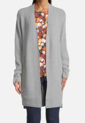 Kobieta ubrana w długi, jasnoszary kardigan zapinany z przodu, nałożony na wielokolorową koszulę z okrągłym wzorem oraz ciemnoniebieskie spodnie.