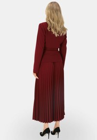 Bordowa spódnica plisowana i dopasowana marynarka, wykonane z teksturowanej tkaniny. Czarne szpilki dopełniają stylizację.