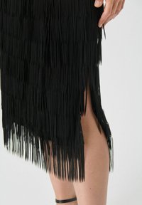 Czarna spódnica z frędzlami o pionowych pasmach, z bocznym rozcięciem. Miękka tekstura i warstwowy design tworzą ruch i kontrast.