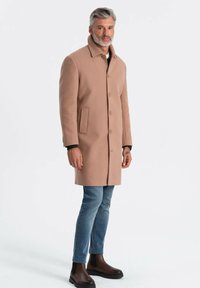 Abrigo de lana beige con cuello, botones en la parte delantera y bolsillos laterales, combinado con vaqueros azules y botas marrones de tobillo.