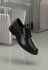 Scarpe eleganti in pelle nera lucida con lacci, elevate sopra un pavimento grigio riflettente vicino a mensole metalliche a rete contenenti libri.