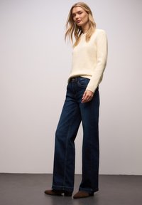 Kremowy sweter z długimi rękawami z dzianiny, zestawiony z ciemnoniebieskimi szerokimi spodniami typu jeans oraz brązowymi botkami na obcasie. Gładka faktura i dopasowany fason.