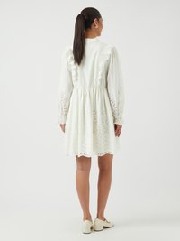 Rochie albă cu umeri volanati, model perforat, și fustă evazată. Mâneci lungi, talie strânsă și lungime până la genunchi, asortată cu încălțăminte ușoară.