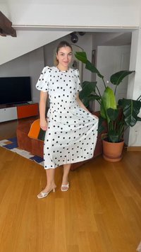 Frau steht in Innenräumen und trägt ein weißes Midikleid mit schwarzen Punkten, hält den Saum, in der Nähe einer großen Topfpflanze und einem Fernsehgerät.