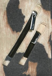 Beige och grå mönstrad tyg med två svarta dragkedjor. Dragkedjehandtagen är metalliska. Tyget har en mjuk textur med ett prickigt mönster.