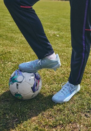 Bottes de football bleu clair avec une tige texturée, posées sur un ballon de football multicolore, sur de l'herbe verte, à côté d'un pantalon de sport bleu marine.