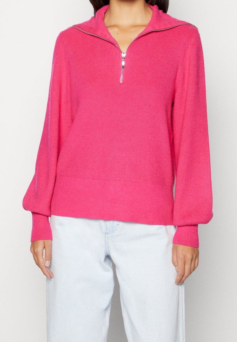 Suéter de punto fucsia brillante con un cuello de media cremallera, textura acanalada y mangas globito, combinado con pantalones de mezclilla azul claro.