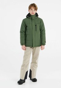 Giacca isolante verde con tasca sul petto con zip, colletto alto e cappuccio nero. Pantaloni beige con dettagli neri ai orli. Stivali neri.