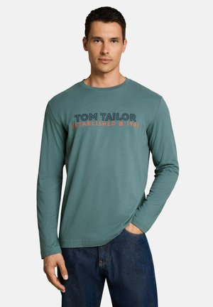 Mann trägt ein türkisfarbenes Langarmshirt mit dem Text "Tom Tailor Established @ 1962" und dunkelblaue Jeans, steht mit einer Hand in der Tasche.