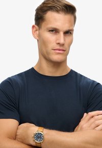 Navy-blaue Baumwoll-T-Shirt mit Rundhalsausschnitt. Person mit einer goldenen und braunen Uhr, Arme verschränkt, neutraler Hintergrund.