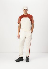 Loungewear komplekts, kas sastāv no krēmkrāsas īsām piedurknēm augšdaļas ar rūsas akcentiem un krēmkrāsas sporta biksēm ar rūsas sānu joslu, kombinēts ar melnā krāsā sneakers.