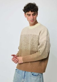 Pull beige et marron clair à motifs avec un col et des poignets côtelés. Le tissu a une texture douce et présente un dégradé de couleurs.