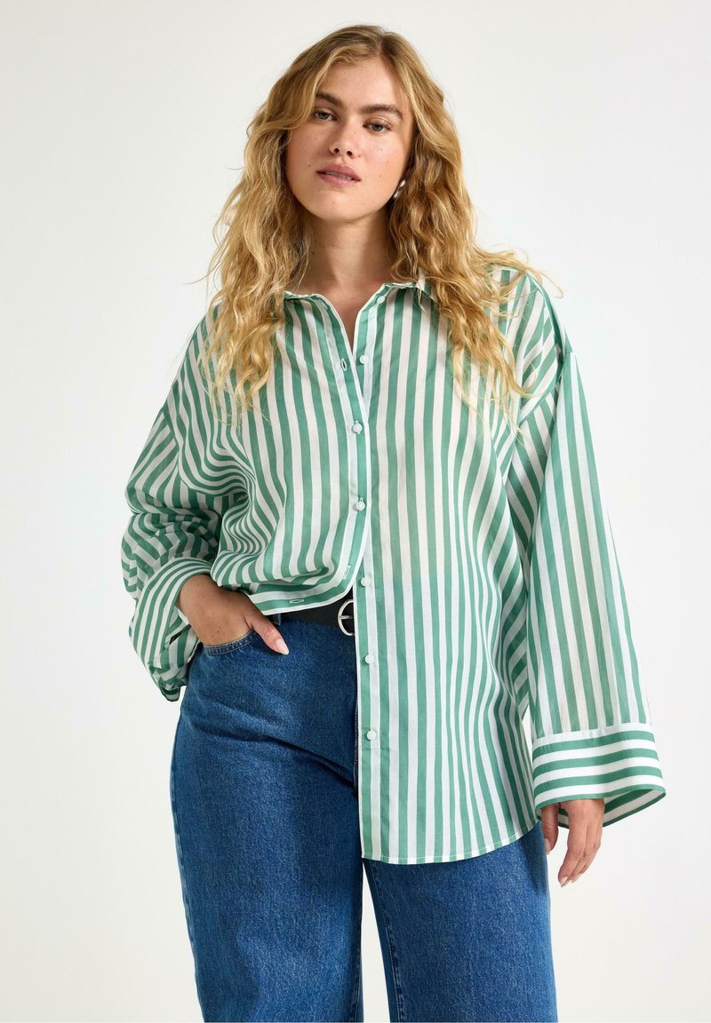 Camicia a righe verdi e bianche con bottoni, maniche lunghe, vestibilità ampia e colletto a punta. Abbinata a pantaloni in denim blu a vita alta.