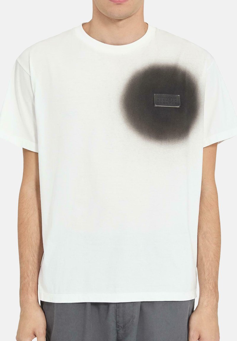 Hombre con camiseta blanca de manga corta con un círculo pintado con spray negro y un pequeño parche rectangular en el pecho izquierdo.