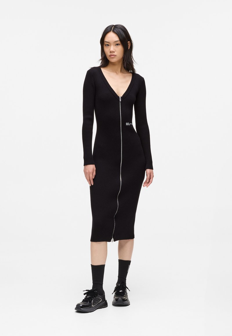 Vestido midi preto canelado com mangas longas e decote em V profundo, apresentando um zíper central e pequeno detalhe com logotipo na lateral.