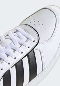 Zapatilla blanca hecha de cuero texturizado con tres franjas negras, puntera redondeada y cordones blancos. Presenta perforaciones para la ventilación.