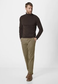 Dark brown turtleneck sweater, slim-fit olive trousers, and brown leather lace-up boots. Model standing with hands in pockets.