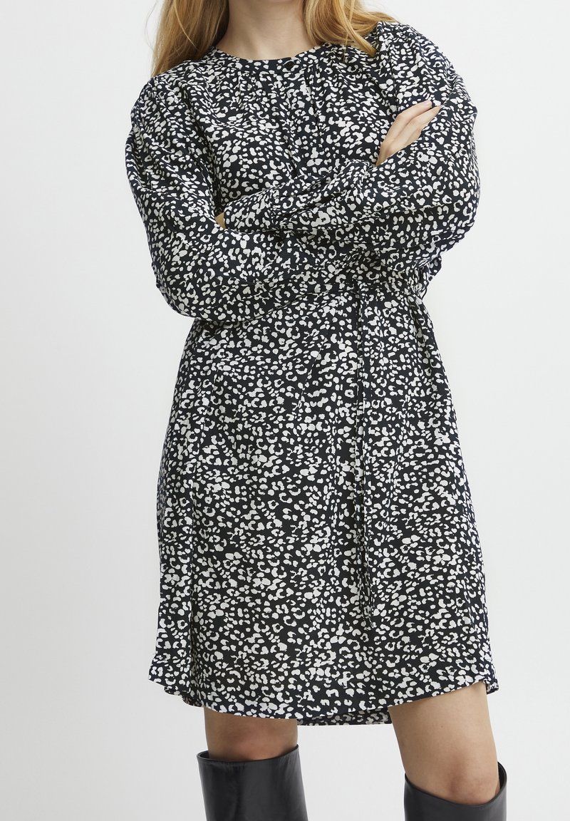 Femme debout, bras croisés, portant une robe noire à taches blanches abstraites et des bottes noires montantes jusqu'aux genoux.