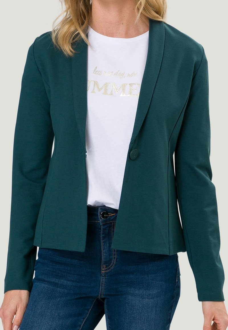 Tyrkysový blazer vyrobený z měkké tkaniny s přizpůsobeným designem, s jedním knoflíkem a dlouhými rukávy, vrstvený přes bílé tričko s potiskem.