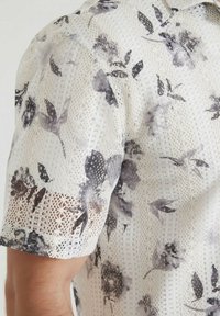 Chemise blanche avec un motif floral en dentelle dans des nuances de gris, présentant une coupe structurée et des manches courtes, mise en valeur par des détails textiles complexes.