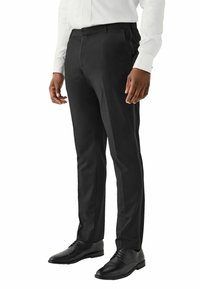 Schwarze Anzughosen mit maßgeschneiderter Passform, aus glattem Stoff gefertigt. Mit flachem Vorderteil, seitlichen Taschen und kombiniert mit klassischen schwarzen Schnürschuhen.