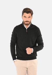 Maglione nero a maglia con colletto a mezza zip, maniche lunghe e orlo a coste. Abbinato a pantaloni chiari.