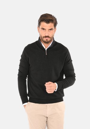 Maglione nero a maglia con colletto a mezza zip, maniche lunghe e orlo a coste. Abbinato a pantaloni chiari.