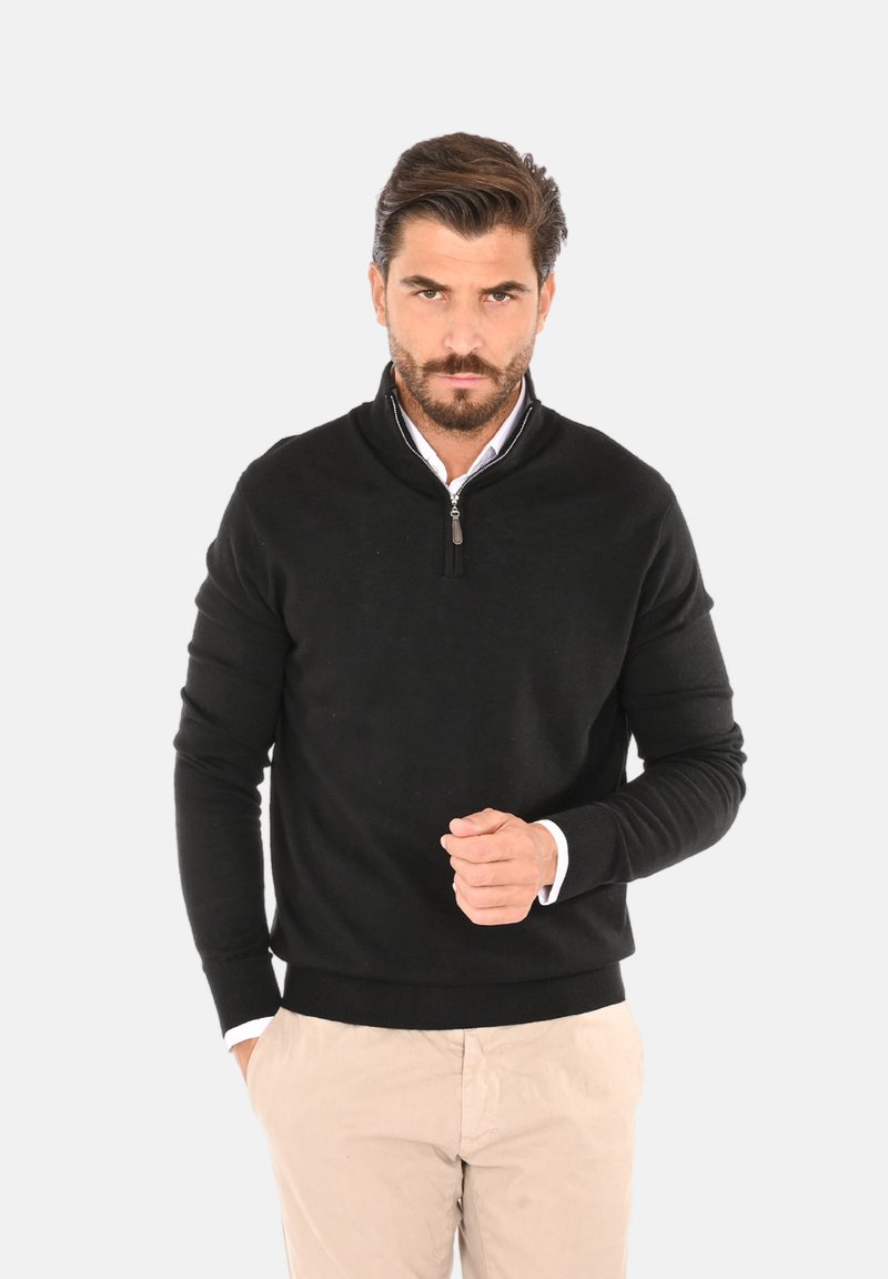 Maglione nero a maglia con colletto a mezza zip, maniche lunghe e orlo a coste. Abbinato a pantaloni chiari.