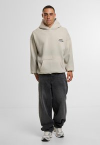 Giovane uomo con capelli corti e lentiggini che indossa un felpa beige con tasca frontale, jeans neri larghi e sneakers bianche in piedi su uno sfondo bianco.