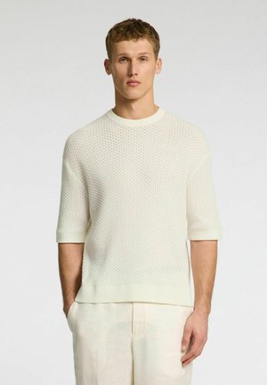 Cremefarbener kurzärmliger Strickpullover mit einem strukturierten Netzdesign, mit rundem Halsausschnitt und gerippten Bündchen, kombiniert mit hellen Hosen.