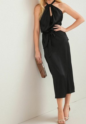 Woman wearing a sleeveless black midi dress with halter neck, holding a brown clutch, and wearing beige high-heeled sandals.
